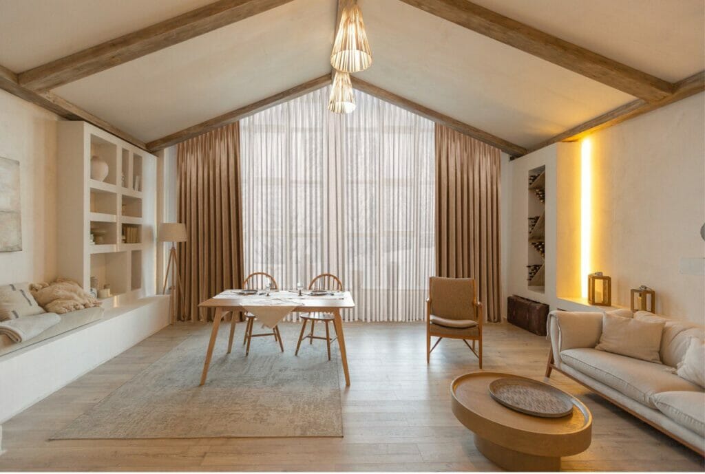A living room with beautiful light curtains, wooden beams and a wooden table.