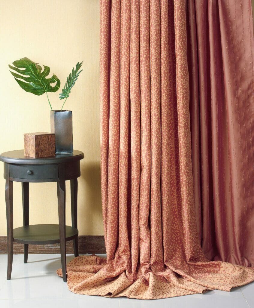 Beige Curtains Pool On The Floor Beside A Dark Wooden Round Table With A Glass Vase Holding Green Leaves.